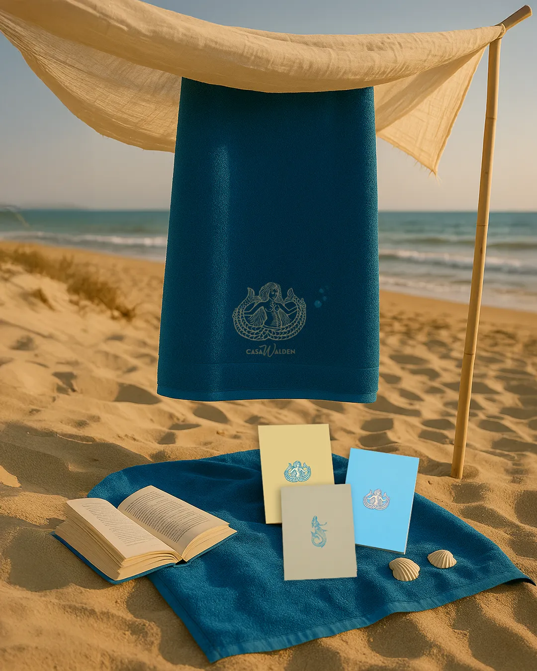 Bundle Sirene 3 Foto ambientata in spiaggia dei prodotti della collezione Sirene by Walden quali: telo mare Ondina azzurro con sirena bicaaudata ricamata a mano e logo Casa Walden, i quaderni Ondina in versione beige e azzurro con illustrazione della serena bicadauta, e quaderno Partenope con copertina in carta martellata beige e icona azzurra di una sirena