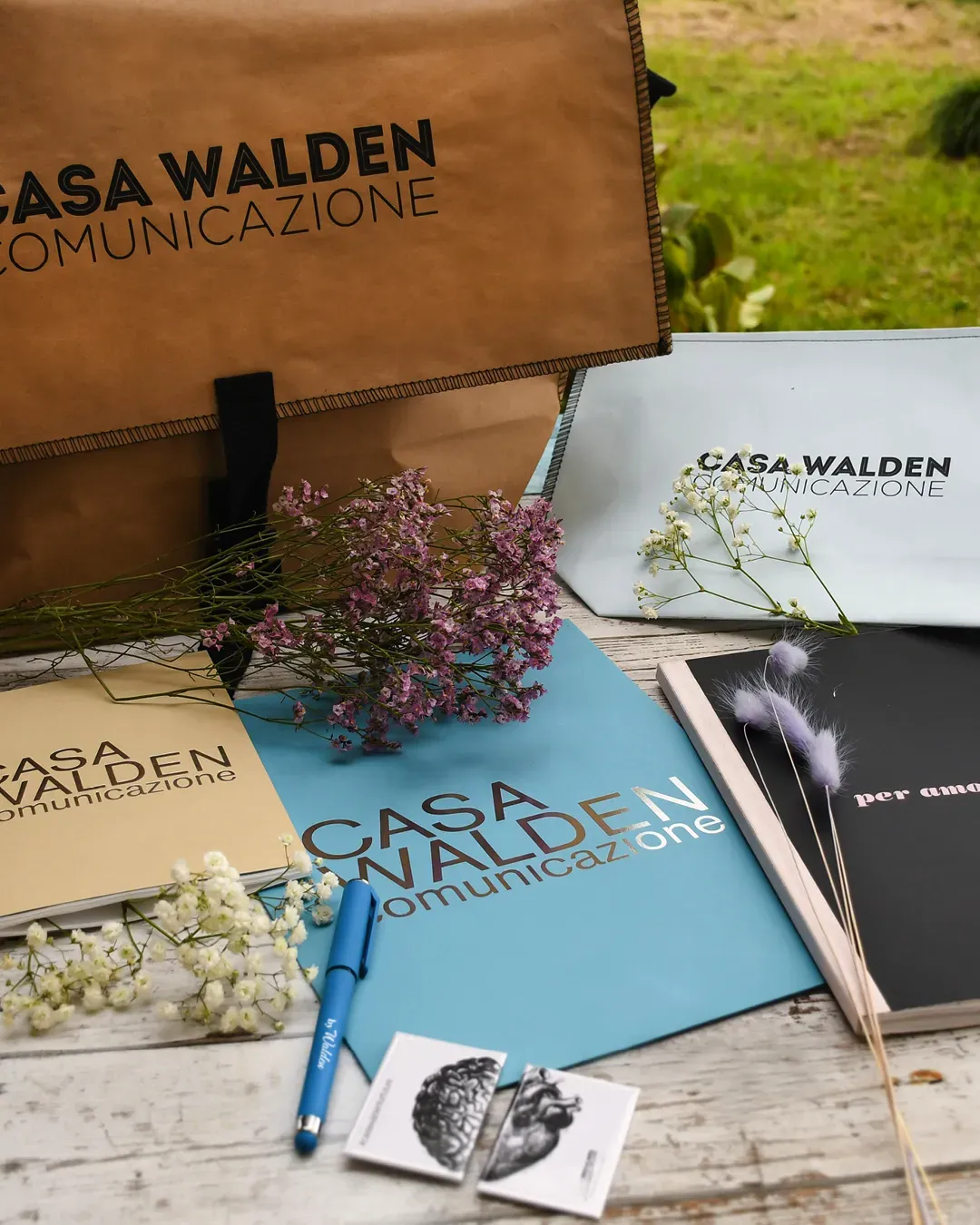 Foto ambientata in antura che mostra la borsa Postina, la pochette, il quaderno, la penna gel, le calamite e il libro monografia dell'agenzia Casa Walden Comunicazione
