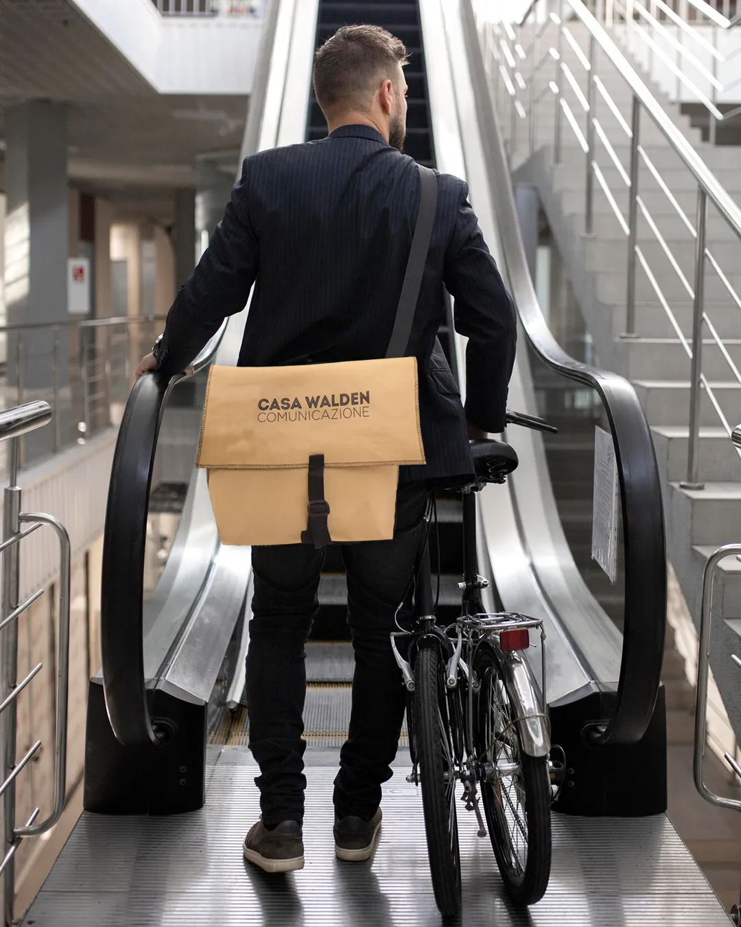 Foto di un uomo ritrattto di spalle, in procinto di salire una scala mobile con la sua bici richiudibile, che indossa la messenger bag La Postina della collezione Walden, realizzata in carta naturale cucita con fibia e tracolla in tessuto nero con al centro il logo di Casa Walden Comunicazione scritto in nero a stampatello.