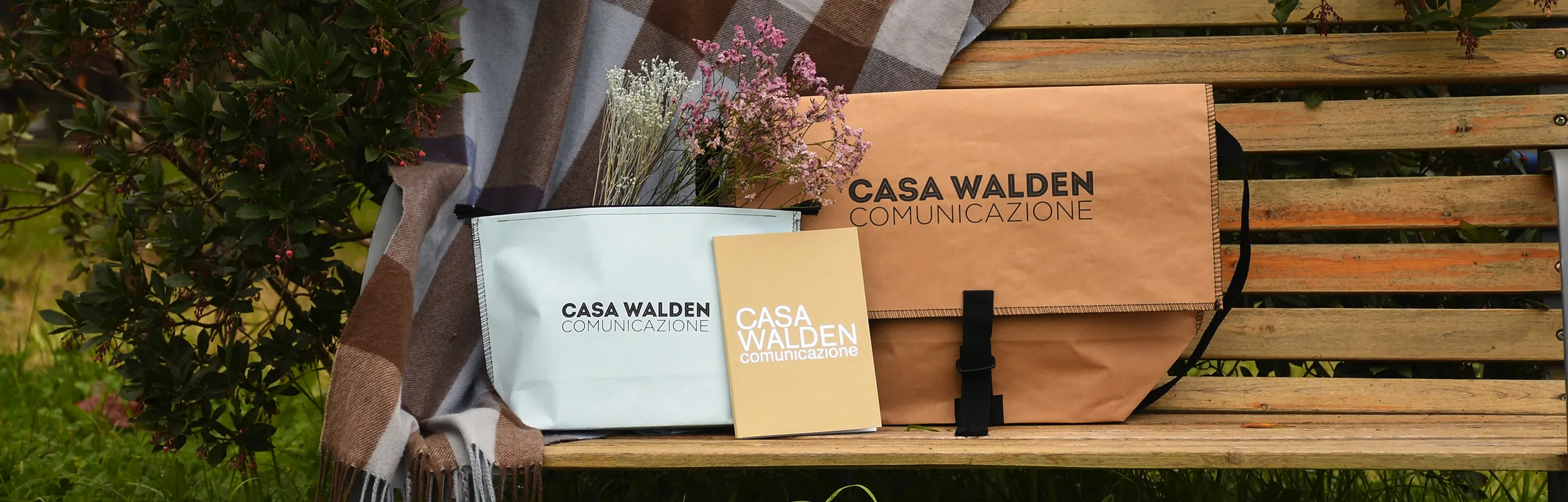 Foto ambientata che mostra la borsa Postina, la pochette e il quaderno brandizzati Casa Walden Comunicazione poggiati su una panchina