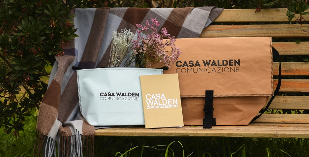 Foto ambientata che mostra la borsa Postina, la pochette, il quaderno e la penna gel brandizzati Casa Walden Comunicazione poggiati su una panchina