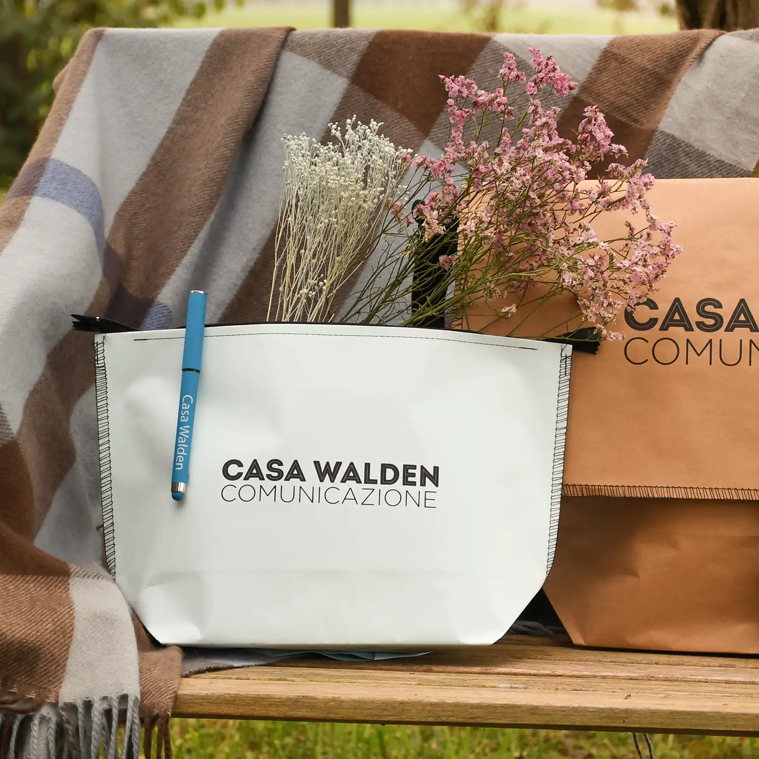 pochette Walden ambientazione Foto ambientata in natura su di una panchina che mostra alcuni prodotti della Collezione Walden tra cui la borsa Postina e la pochette di carta, con la penna gel, brandizzati Casa Walden Comunicazione