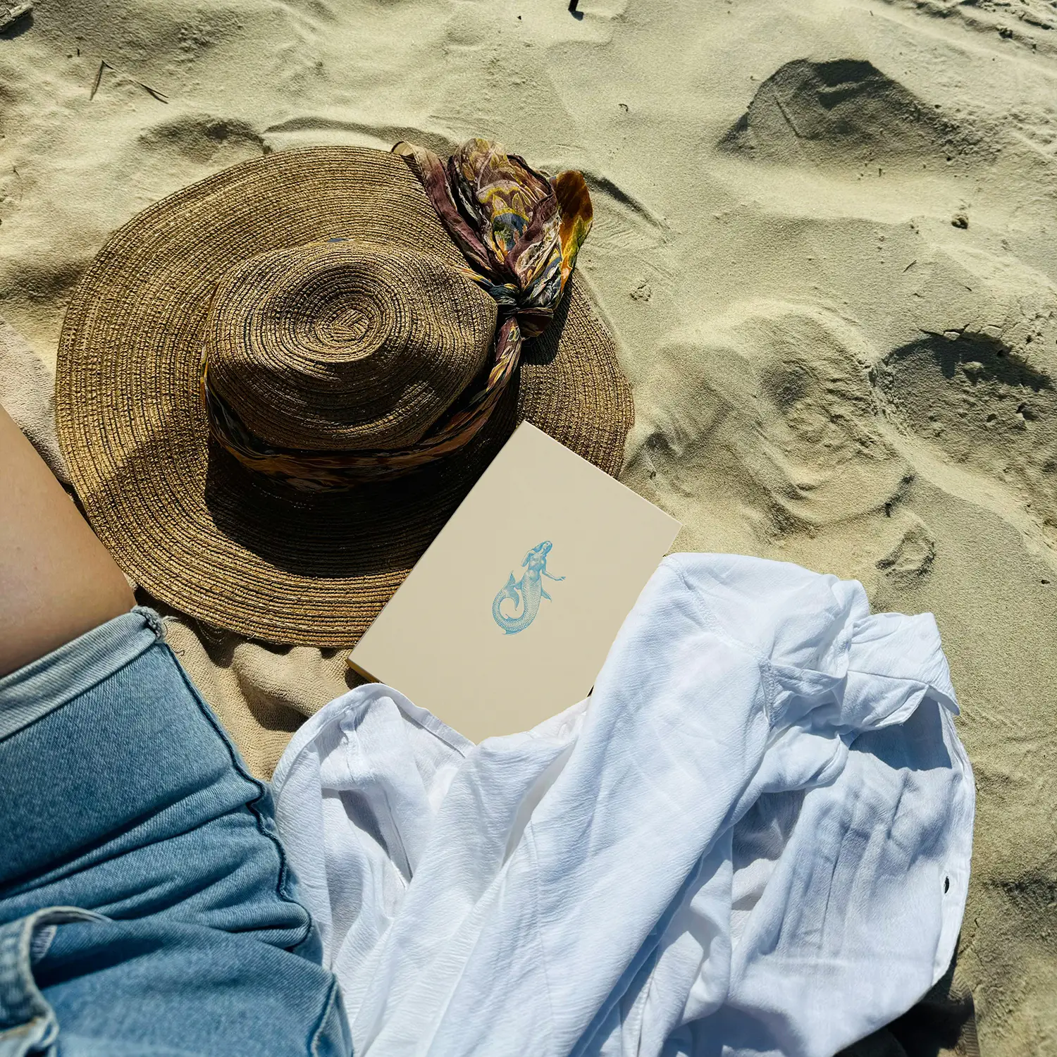quaderno A5 Sirene Partenope ambientazione Foto ambientata in spiaggia del Quaderno Partenope con copertina in carta martellata beige e icona azzurra di una sirena