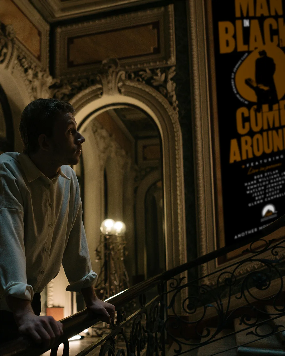 Uomo in camicia bianca, appoggiato sulla ringhiera di una scala, guarda verso l’alto, dove su una parete interna decorata è appeso un grande poster cinematografico nero e arancione con la scritta “BLACK COMES AROUND” e la sagoma di un uomo con cappello.