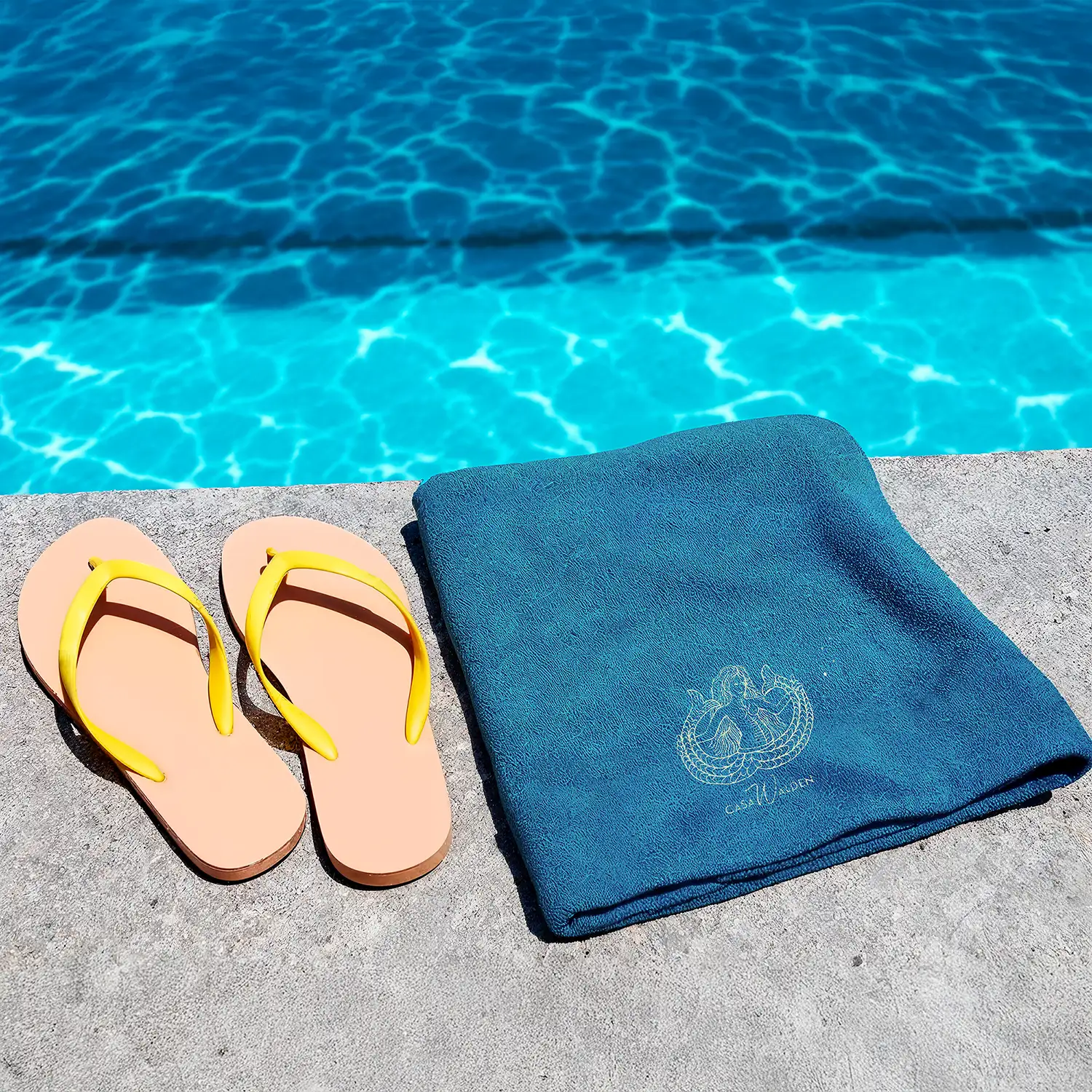 Telo Marea, ambientazione Foto ambientata a bordo piscina di un paio di infradito con fascia gialla con accanto il Telo mare Marea, piegato, di color azzurro con ricamo in evidenza di una sirena bicaudata, tre bollicine e logo di Casa Walden.