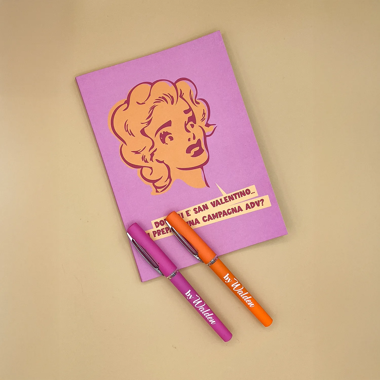 collezione 1950s, quaderno Dolly con Penne Due penne gel di colore fucsia e arancio, con logo bianco 'byWalden', tappo protettivo con finestrella trasparente e clip metallica per aggancio, poggiate sul quaderno Dolly dalla copertina rosa illustrata con testa cartoon anni ’50 e slogan pubblicitario ‘Domani è San Valentino, mi prepari una campagna ADV?’